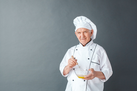 Senior chef studio standing isolated on gray holding whisk mixing eggs looking camera playfulの写真素材