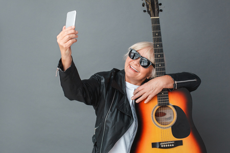 Senior woman in leather jacket and sunglasses studio standing isolated on gray with guitar taking selfie on smartphone happyの写真素材