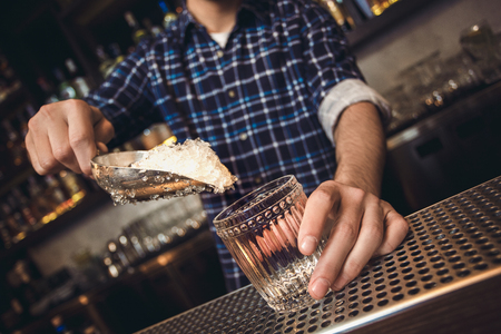 Young bartender standing at bar counter putting shaved ice into glass close-upの写真素材