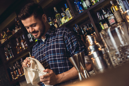 Young bartender standing at bar counter wiping glass smiling cheerfulの写真素材