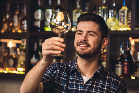 Young bartender standing at bar counter looking at wine glass dreamfulの写真素材