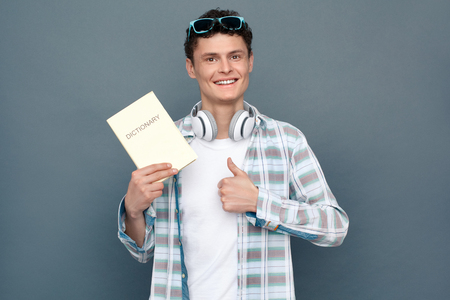Man isolated on gray wall tourism concept standing with dictionary showing thumb up cheerfulの写真素材
