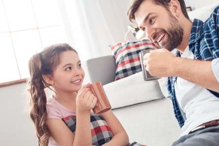 Father and little daughter at home sitting on floor drinking hot tea smiling playfulの写真素材
