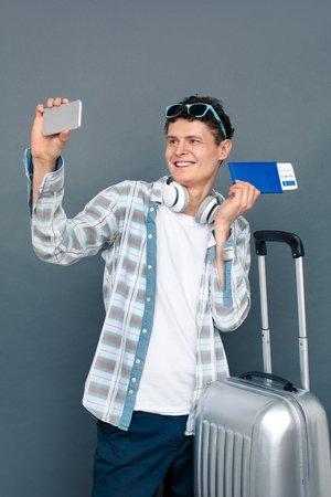 Man isolated on gray wall tourism concept standing with suitcase holding passport and ticket taking selfies on smartphone looking camera cheerfulの写真素材