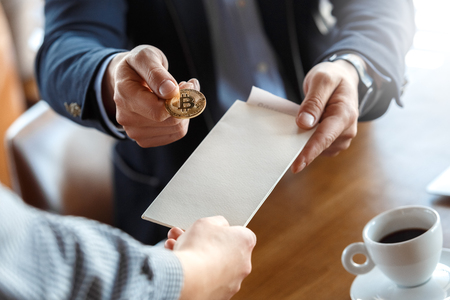 Mature man trader sitting at table at cafe with cup of hot coffee taking bill paying to waiter with cryptocurrency coin interaction close-upの写真素材