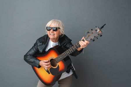 Senior woman in leather jacket and sunglasses studio standing isolated on gray playing guitar cheerfulの写真素材