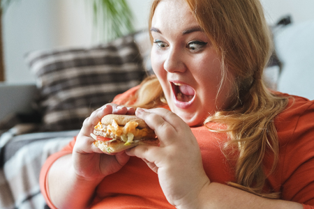 Chubby woman sitting at home ready to eat burger excitedの写真素材