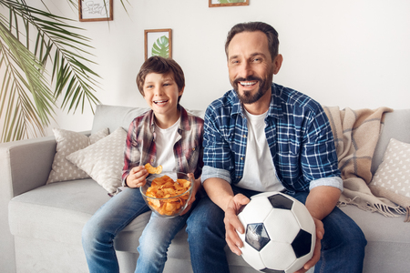 Father and little son at home sitting on sofa boy eating potato chip dad with ball watching football championship cheerfulの写真素材