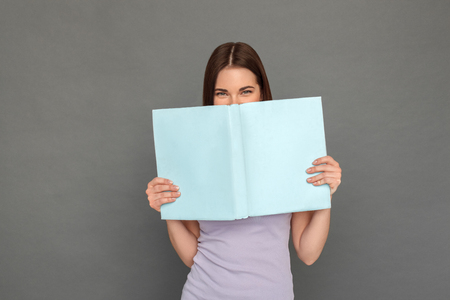 Freestyle. Young girl standing isolated on grey covering face with book smiling playfulの写真素材