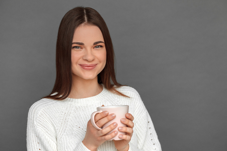 Freestyle. Young girl standing isolated on grey drinking hot tea looking camera cheerful close-upの写真素材