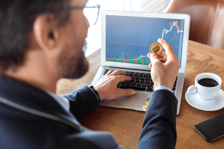 Business Lifestyle. Trader sitting at cafe with coffee checking trading chart on laptop holding cryptocurrency coin back view close-upの写真素材