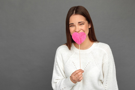 Freestyle. Young girl standing isolated on grey with heart on stick covering mouth laughing playfulの写真素材