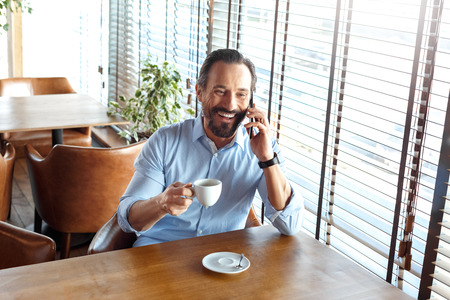 Business Lifestyle. Trader sitting at cafe drinking hot coffee talking on smartphone laughing cheerfulの写真素材