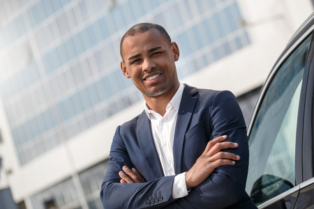 Young businessman standing near car crossed arms looking camera cheerful close-upの写真素材