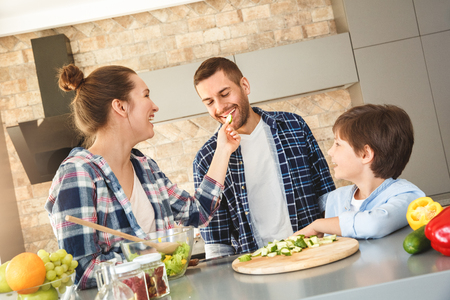 Family at home standing in kitchen together wife feeding husband with cucumber slice laughingの写真素材