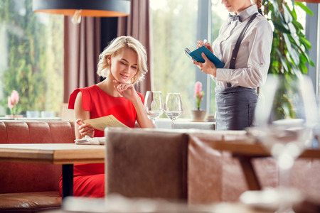 Young woman dining in restaurant sitting choosing dish while waiter waiting for orderの写真素材