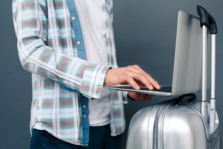 Man isolated on gray wall tourism concept standing with suitcase typing message on laptop close-upの写真素材
