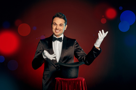 Professional Occupation. Magician in suit and gloves standing isolated on wall making trick with hat on table showing result smiling cheerfulの写真素材