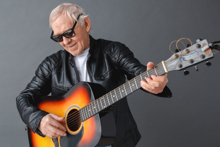 Senior man in leather jacket and sunglasses standing isolated on gray playing guitar smiling joyful close-upの写真素材