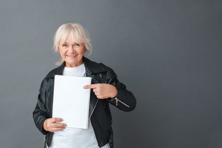 Senior woman in leather jacket studio standing isolated on gray with notebook pointing at paper smiling happyの写真素材