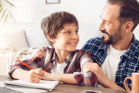 Father and son at home sitting at table dad hugging boy looking at each other happy while doing homework close-upの写真素材