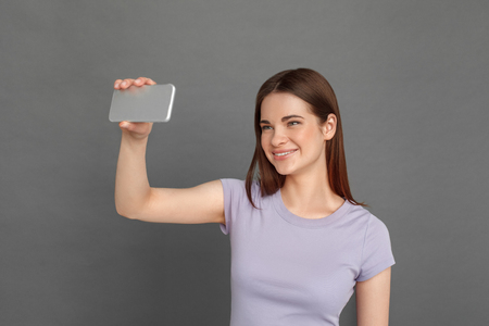 Freestyle. Young girl standing isolated on grey taking selfie on smartphone smiling playfulの写真素材