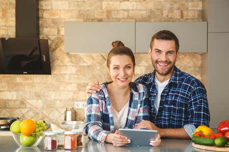 Young couple at home standing in kitchen together husband hugging wife holding digital tablet looking camera happyの写真素材