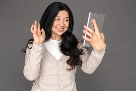 Freestyle. Mature lady standing isolated on grey having video call waving to camera friendlyの写真素材