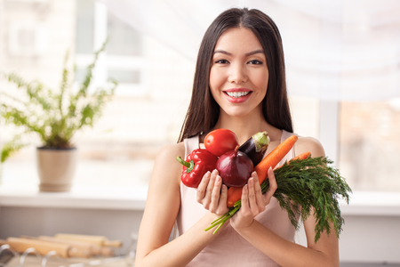 Young girl at kitchen standing holding vegetables looking camera joyful promoting healthy lifestyleの写真素材
