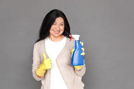 Freestyle. Mature lady in rubber gloves standing isolated on grey with spray thumb up smiling happyの写真素材