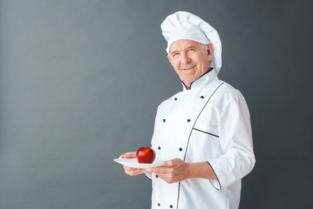 Senior chef studio standing isolated on gray with apple on dish looking camera cheerfulの写真素材