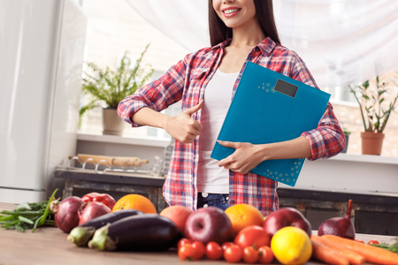 Young girl at kitchen healthy lifestyle standing near table holding scales cheerful showing thub up close-upの写真素材