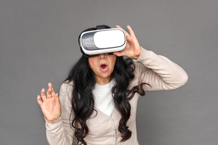 Freestyle. Mature lady in virtual reality headset standing isolated on grey playing surprisedの写真素材