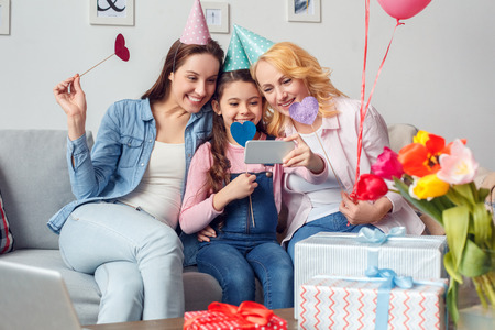 Grandmother mother and daughter together at home celebration sitting in festive caps hugging taking selfie photos with paper heartsの写真素材