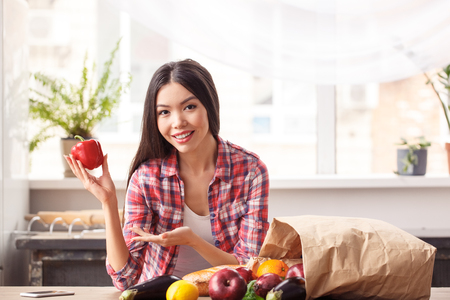 Young girl at kitchen healthy lifestyle standing leaning on table showing bell pepper to camera smilingの写真素材