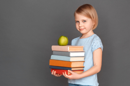 Good student. Little girl isolated on grey with pile of books and apple smiling modestの写真素材