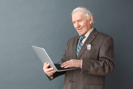 Senior man studio standing isolated on gray browsing internet on laptopの写真素材