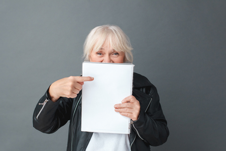 Senior woman in leather jacket studio standing isolated on gray with notebook in front pointing at paper cheerful close-upの写真素材