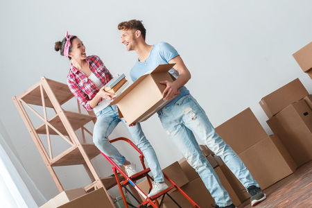 Young couple moving to new place woman standing on ladder putting books into box laughing cheerful with manの写真素材