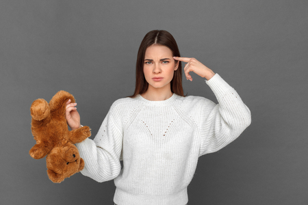 Freestyle. Young girl standing isolated on grey with teddy bear pointing finger gun at head irritatedの写真素材