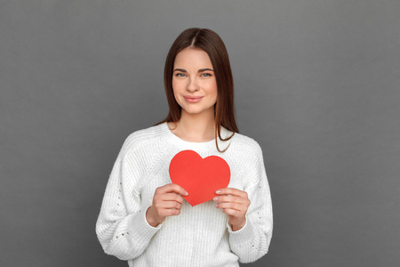 Freestyle. Young girl standing isolated on grey with crafted heart card smiling happyの写真素材