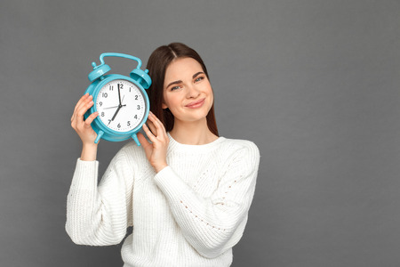 Freestyle. Young girl standing isolated on grey with clock listening to ticking smiling cheerfulの写真素材