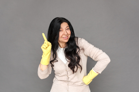 Freestyle. Mature lady in rubber gloves standing isolated on grey pointing aside looking camera playfulの写真素材