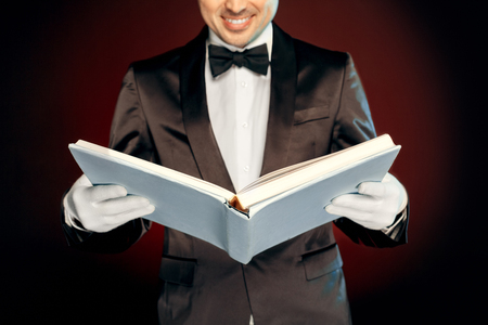 Professional Occupation. Magician in suit and gloves standing isolated on wall reading magic book joyful close-upの写真素材