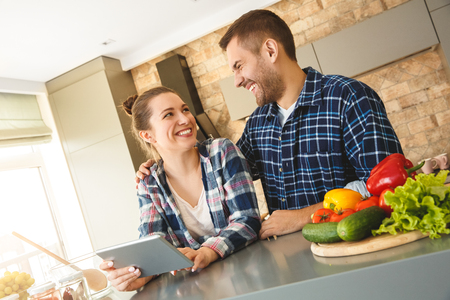 Young couple at home standing in kitchen together husband hugging wife using digital tablet looking at each other laughingの写真素材