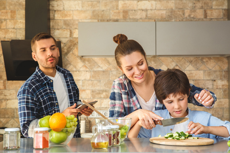 Family at home standing in kitchen together mother teaching son cutting cucumber joyful while father with digital tablet looking at them curiousの写真素材