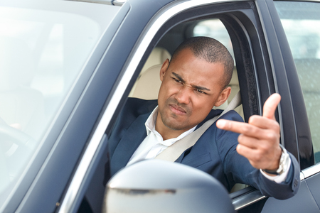 Young businessman driver sitting inside car driving showing middle finger outside offended to another vehicle side view close-upの写真素材