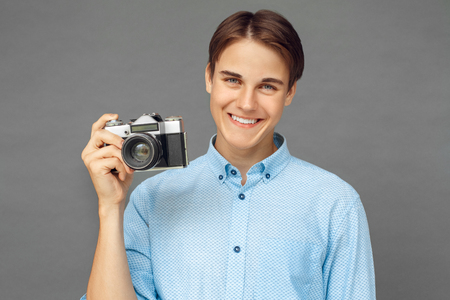 Photography hobby. Teenager boy standing isolated on grey with film camera smiling cheerful close-upの写真素材