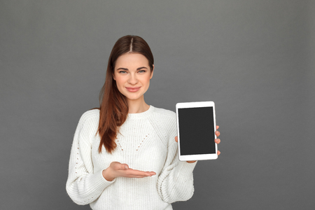 Freestyle. Young girl standing isolated on grey showing screen of digital tablet smiling friendlyの写真素材