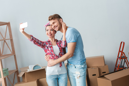 Young couple moving to new place standing hugging taking selfie photos with smartphone smiling joyfulの写真素材